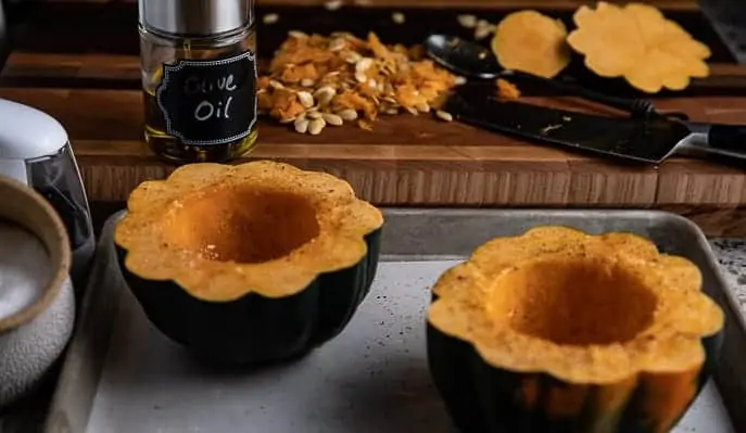 acorn squash cut in half on a baking sheet with parchment paper, olive oil, salt, seeds, knife in background