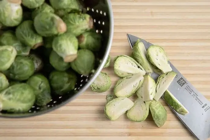 raw brussels sprouts in a metal colander on a wooden board with cut brussels sprouts and a knife