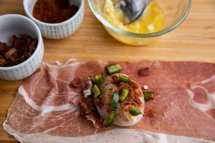 slice of prosciutto on a wooden board with a scallop covered with chopped jalapeno,bacon bits and chipotle seasoning; butter in a dish, chipotle seasoning in a dish and bacon bits in a dish nearby