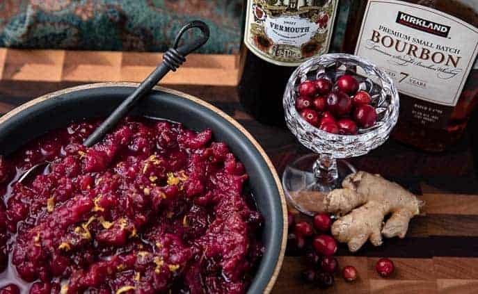 bowl with Instant Pot Manhattan Cranberry Sauce, martini glass with cranberries, bottle of bourbon and sweet vermouth, fresh piece of ginger root and fresh cranberries on a wooden board
