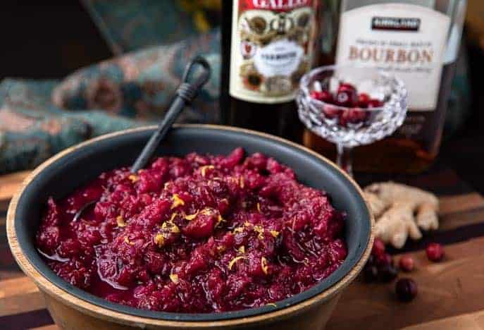 bowl with Instant Pot Manhattan Cranberry Sauce, martini glass with cranberries, bottle of bourbon and sweet vermouth, fresh piece of ginger root and fresh cranberries on a wooden board