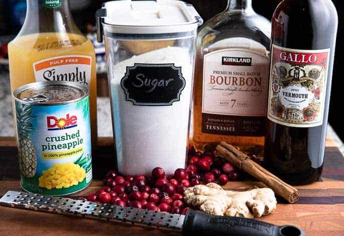 orange juice, can of pineapple, sugar, bourbon, sweet vermouth, cranberries, cinnamon, ginger and grater on a wooden board