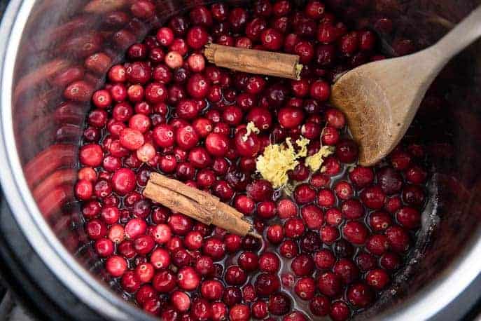 Instant Pot with cranberries, sticks of cinnamon and grated ginger with a wooden spoon
