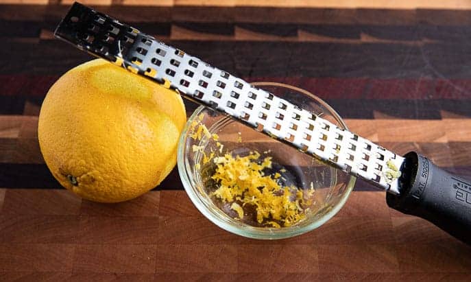 grated orange zest in a glass bowl with orange and grater on a wooden board