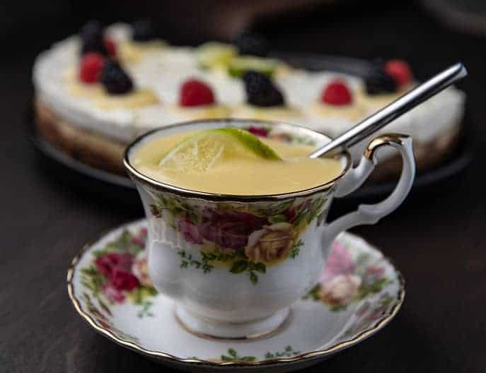 lime curd in a tea cup with piece of lime and spoon; cheesecake with berries in background