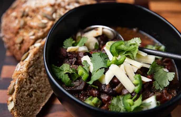 Healthy Soup Recipes: Caribbean Black Bean Soup in a black bowl with a spoon, cilantro, jalapeno on top; bread in background on a wooden board