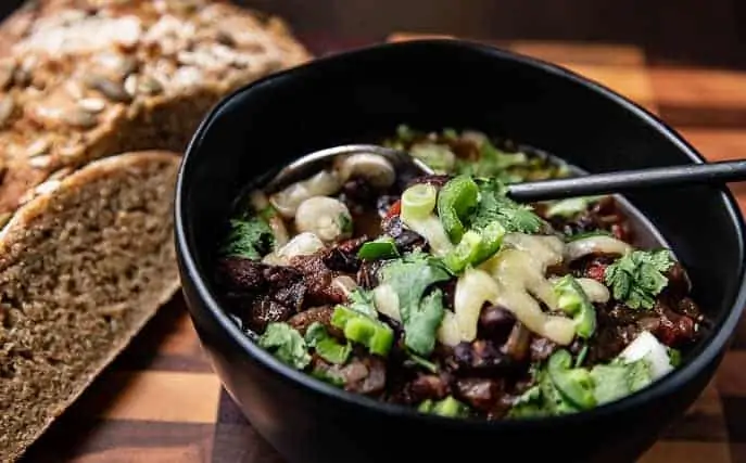 Healthy Soup Recipes: Caribbean Black Bean Soup in a black bowl with a spoon, cilantro, jalapeno on top; bread in background on a wooden board