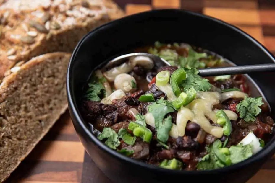 Healthy Soup Recipes: Caribbean Black Bean Soup in a black bowl with a spoon, cilantro, jalapeno on top; bread in background on a wooden board