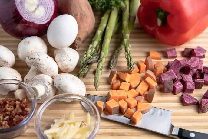 mushrooms, red bell pepper, sweet potatoes, bacon, cheese, egg, asparagus all on a wooden cutting board with knife