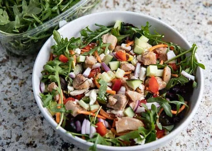 healthy greens with chicken, red pepper, onion, cucumber, avocado in a white bowl on a granite counter