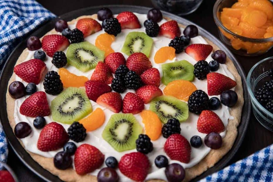 fruit pizza on a table with bowls of strawberries, blueberries, mandarins and blackberries with a blue checked napkin