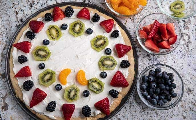 partially finished fruit pizza on a table with bowls of strawberries, blueberries, mandarins and blackberries on a granite counter