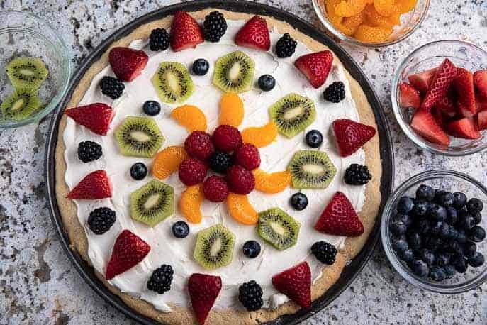 fruit pizza on a table with bowls of strawberries, blueberries, mandarins and blackberries on a granite counter