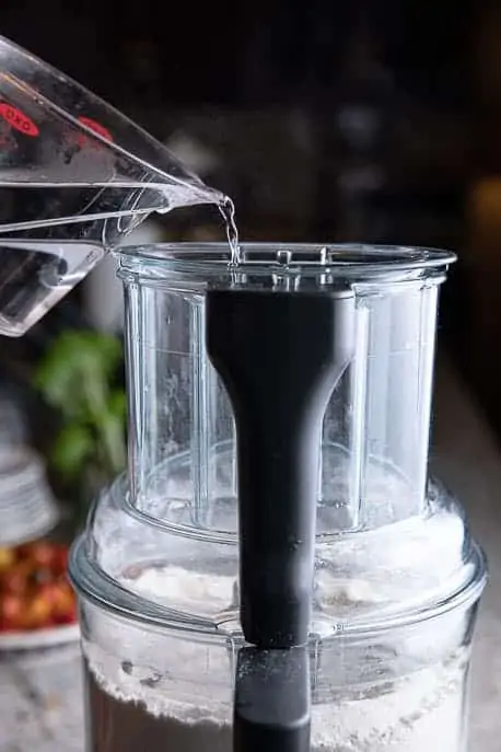 measuring cup pouring water into a food processor with flour mixture