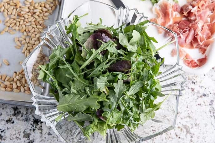 arugula in a glass bowl with toasted pine nuts and prosciutto on the side