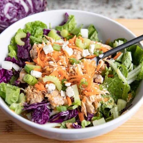 white bowl with black spoon on a wooden board with buffalo chicken, cabbage, celery, blue cheese, green onions, cabbage and carrots